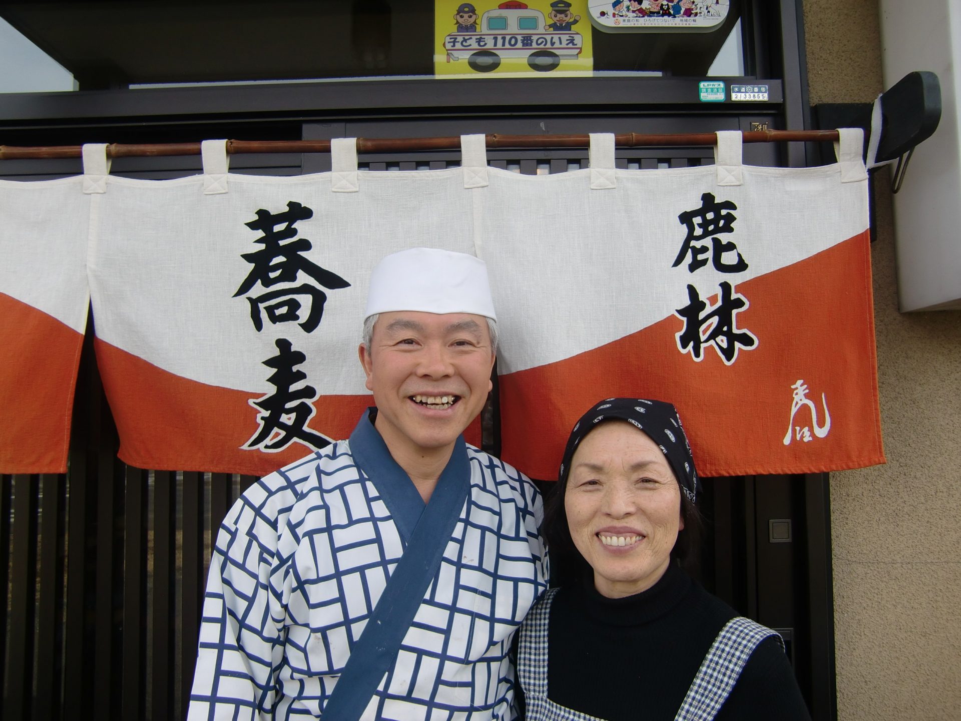札幌市南区豊滝在住 豊野様ご夫妻。そば店の暖簾の前で